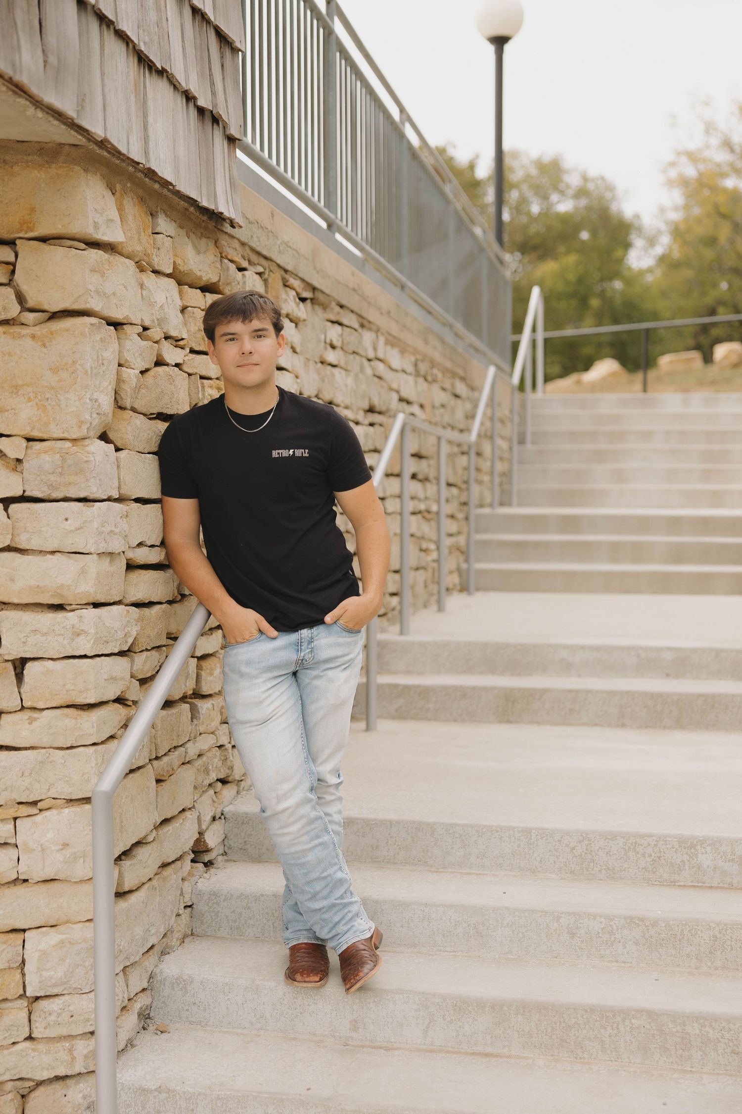 Person in casual attire leans against stone wall by concrete stairs with metal railing and lamp post.