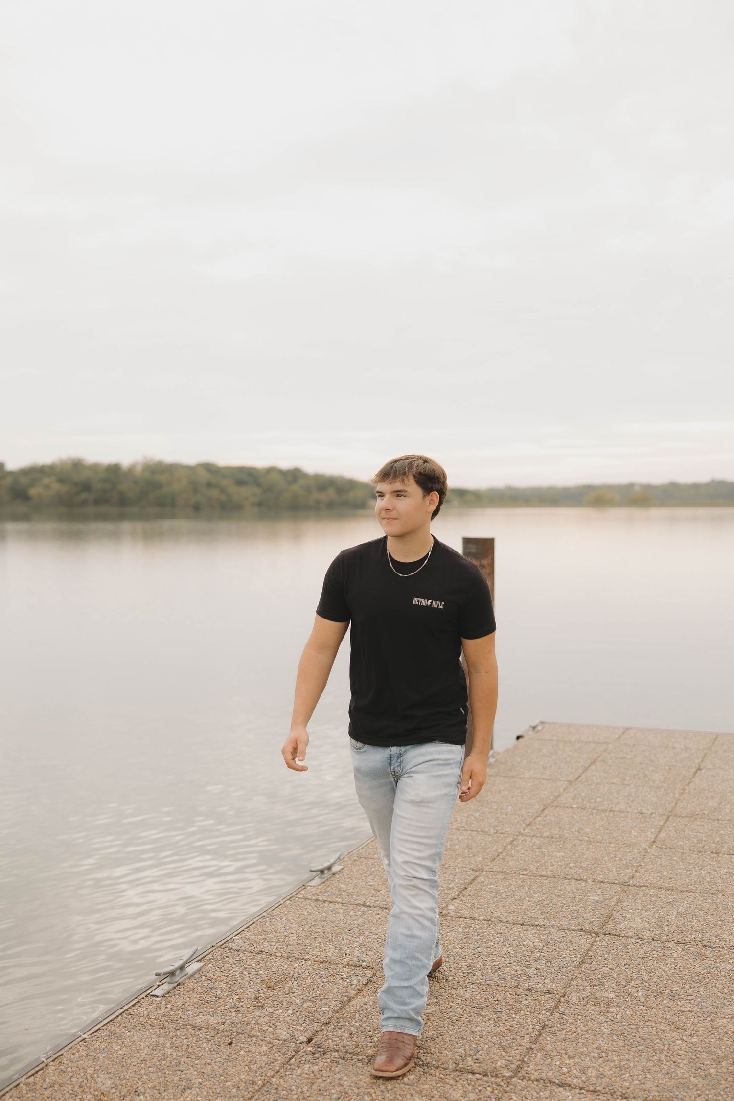 Person in black t-shirt and jeans walks along concrete waterfront path beside calm lake on cloudy day.