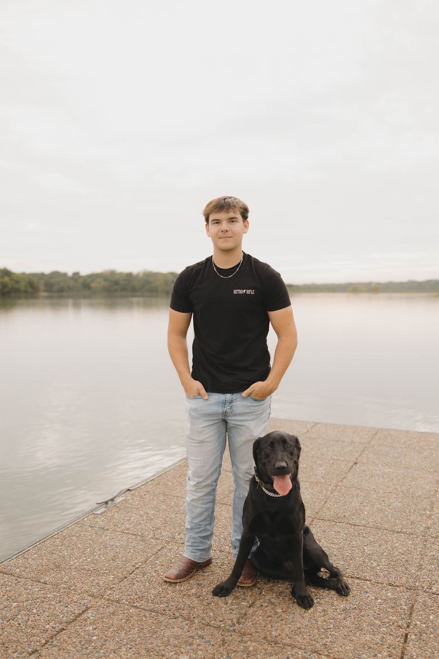 A person in a black t-shirt stands with their black Labrador dog on a concrete pier by a calm lake.