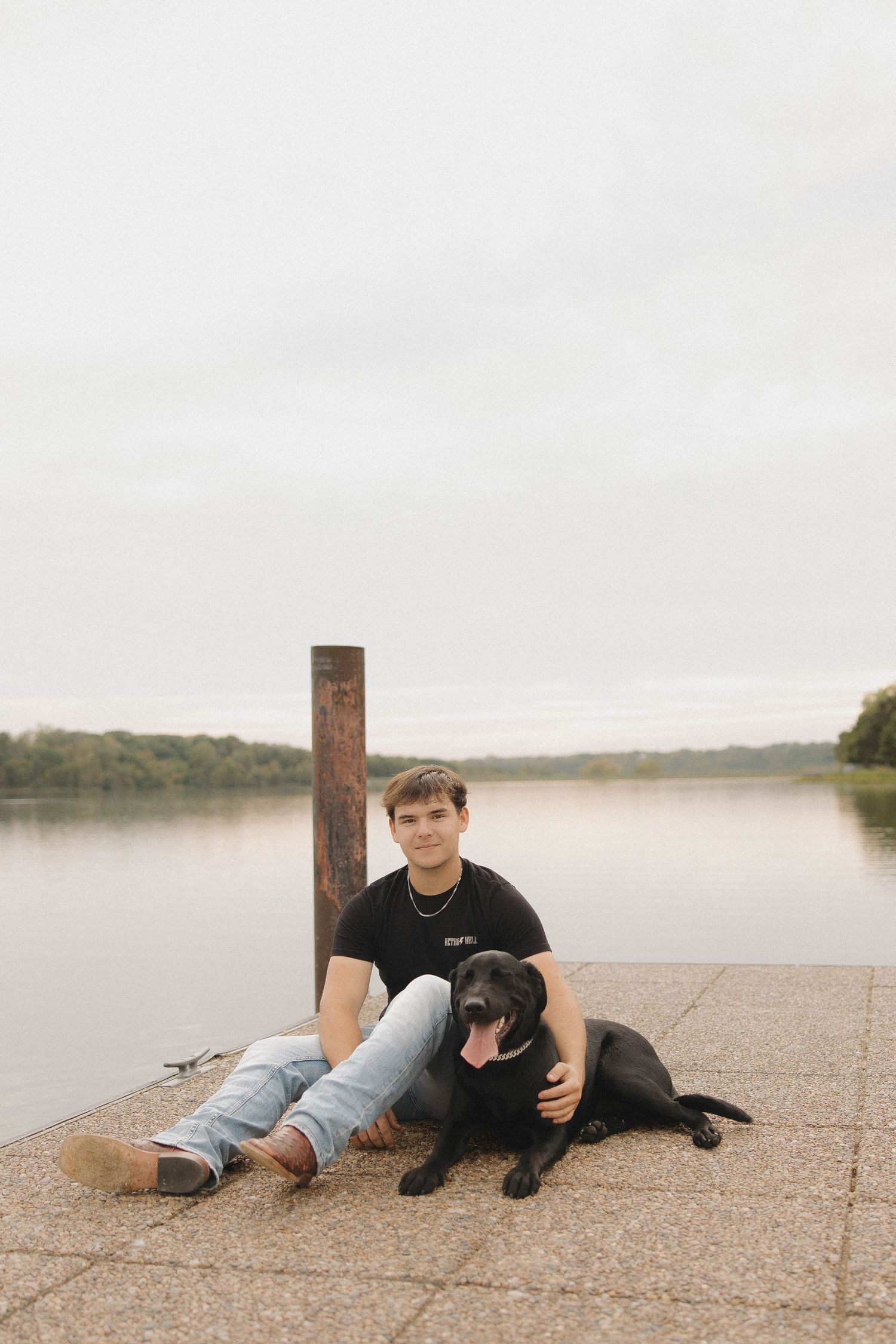 Person relaxing on a beach with a black dog by a lake at sunset.
