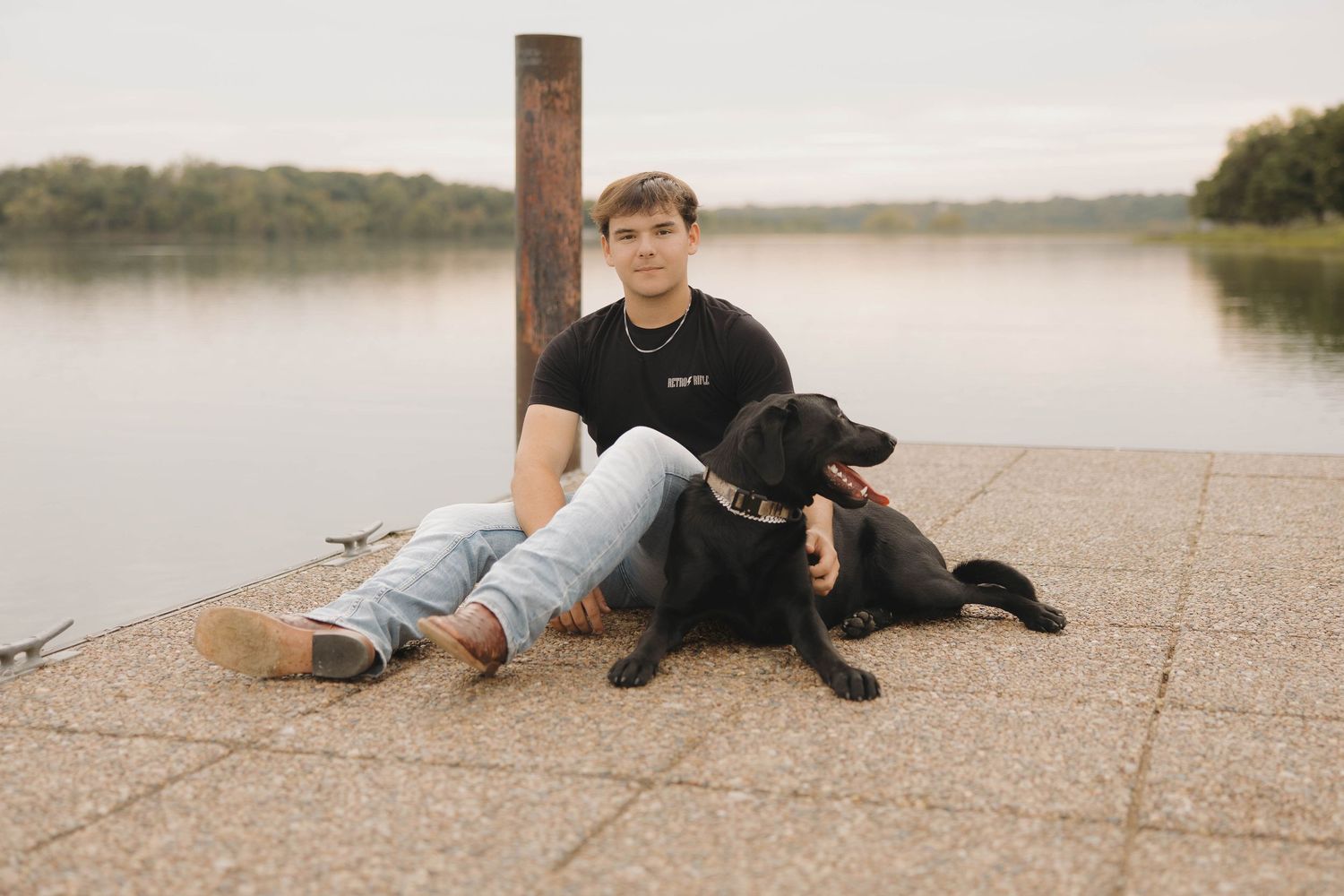 A black dog and their companion sit together on a dock overlooking a serene lake at sunset.