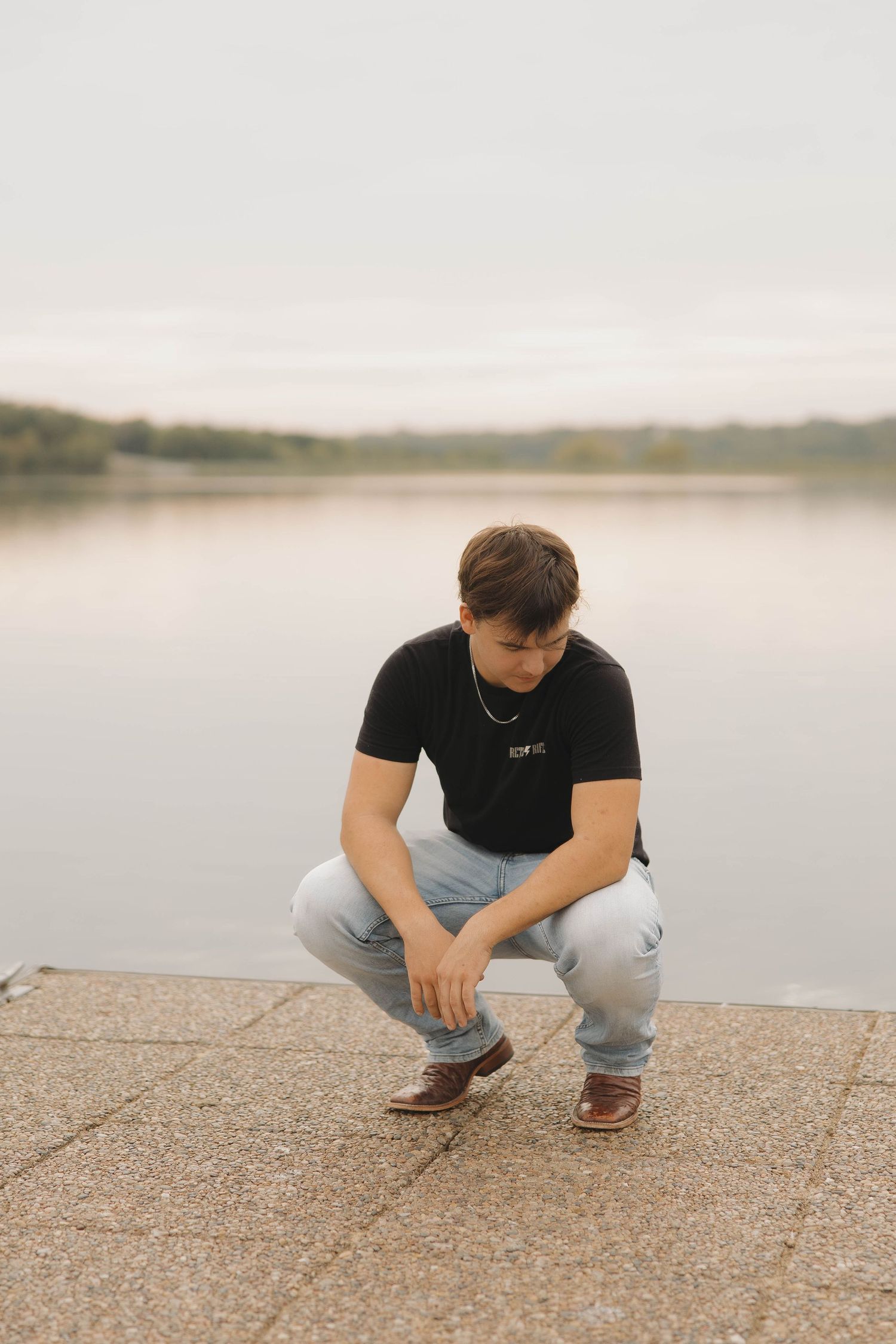 Person in black t-shirt and gray pants crouches down near a serene lakeside on a cloudy day.