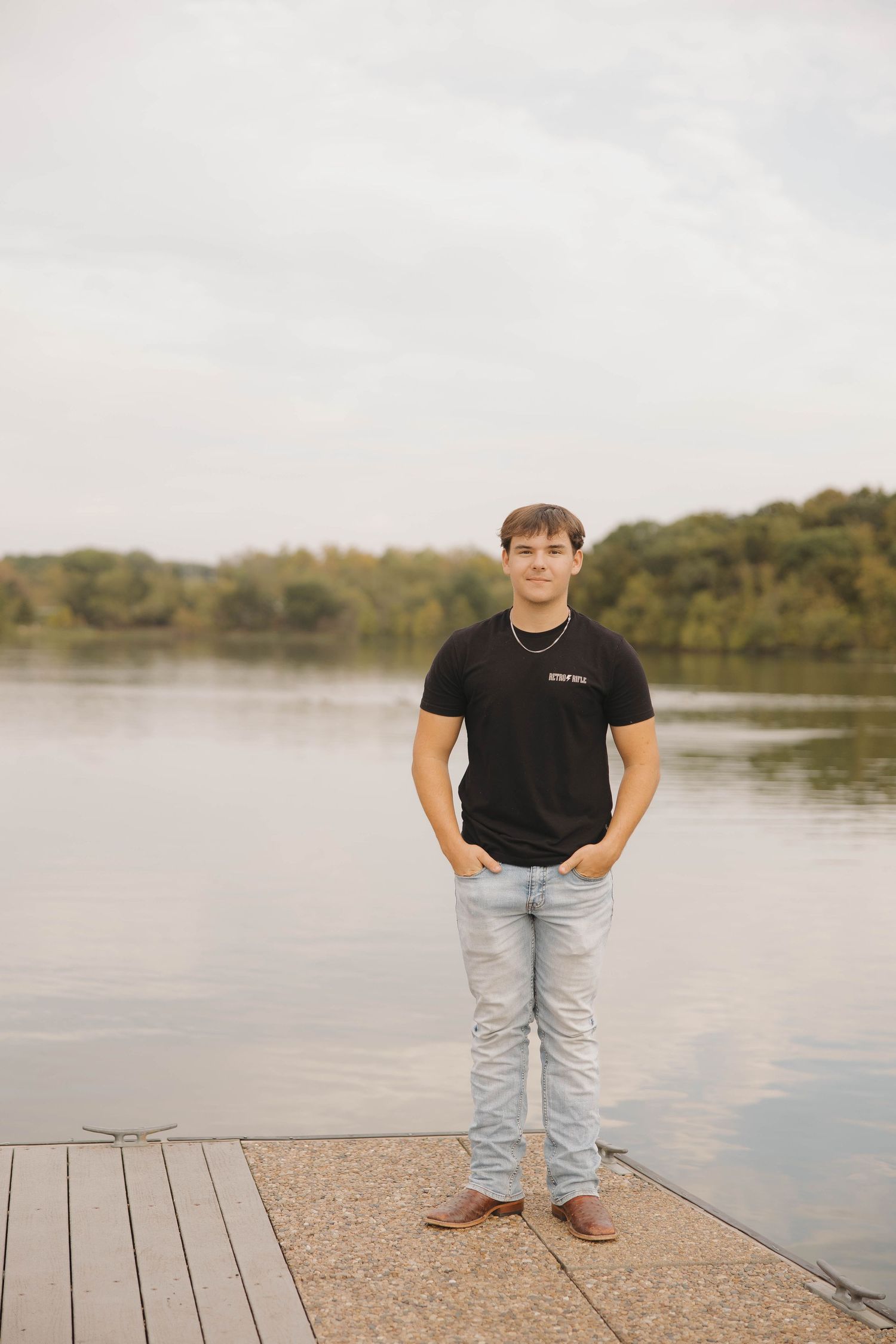 A person in casual attire poses on a wooden dock overlooking a serene lake with autumn trees in the background.