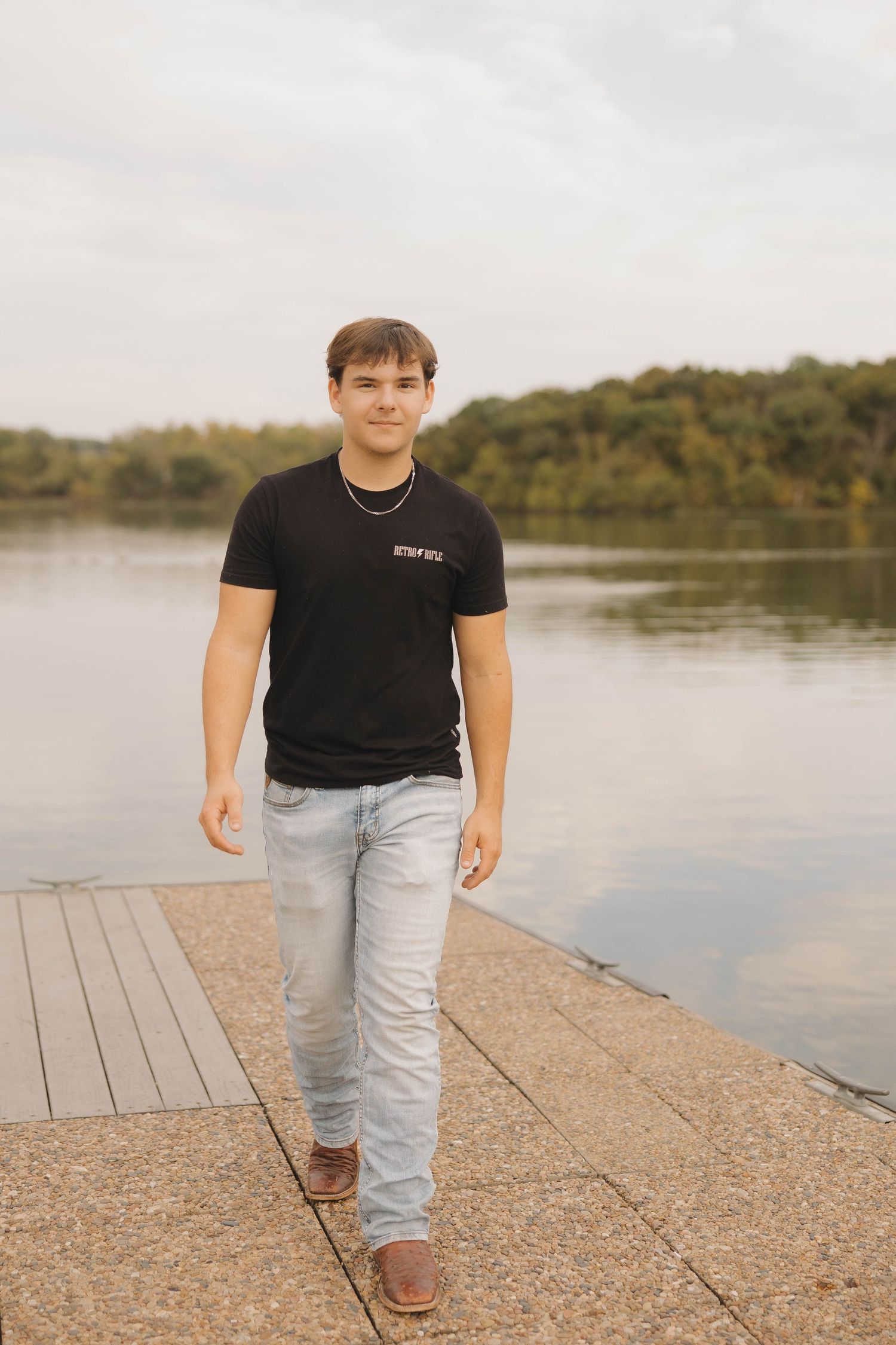 Person wearing black t-shirt and light jeans stands on wooden dock by tranquil lake.