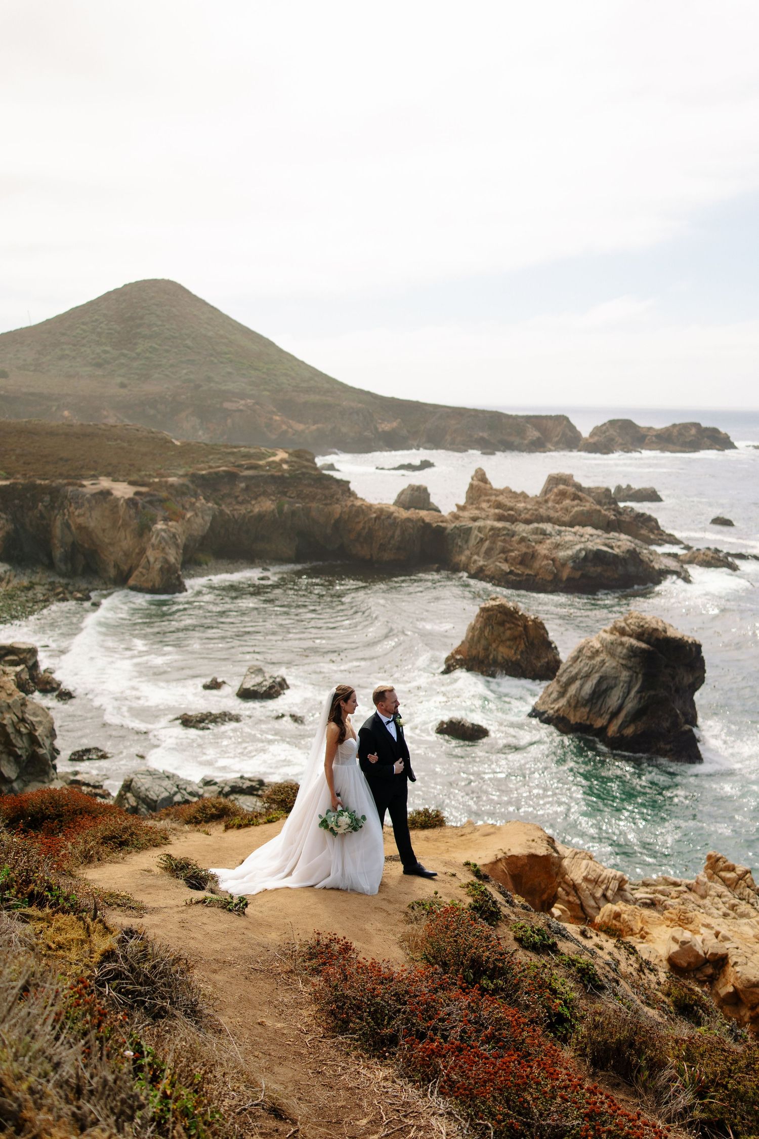 A couple in wedding attire stands on a dramatic coastal cliff overlooking crashing waves and rocky shoreline at sunset.