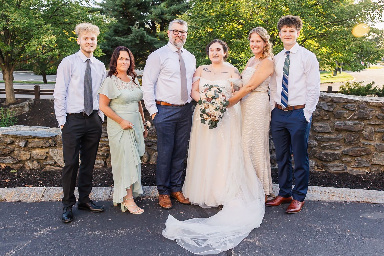 Wedding party poses outdoors in formal attire near stone wall during summer celebration.