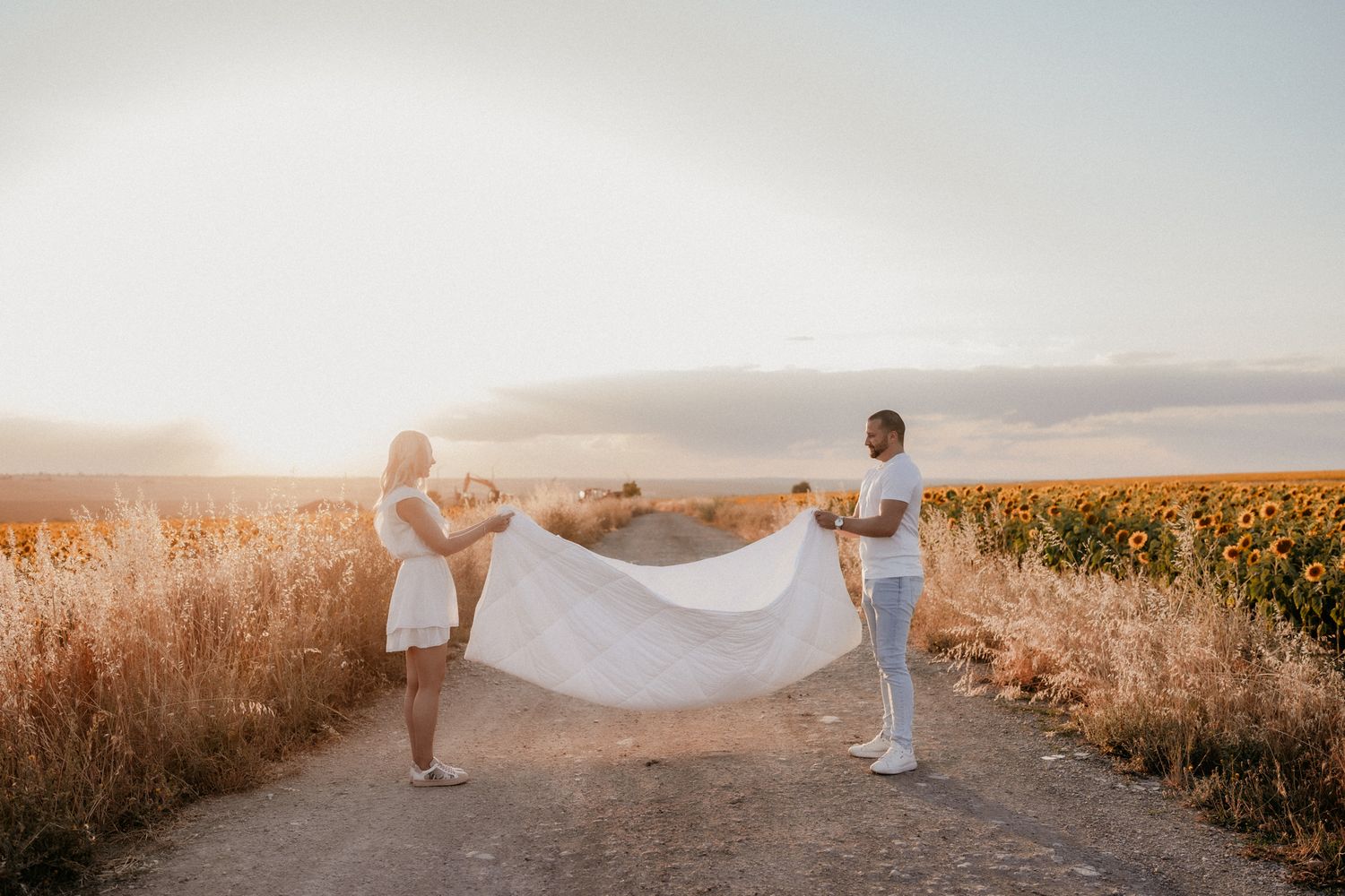 Couple in white outfits holding flowing fabric in a sunflower field during golden hour.