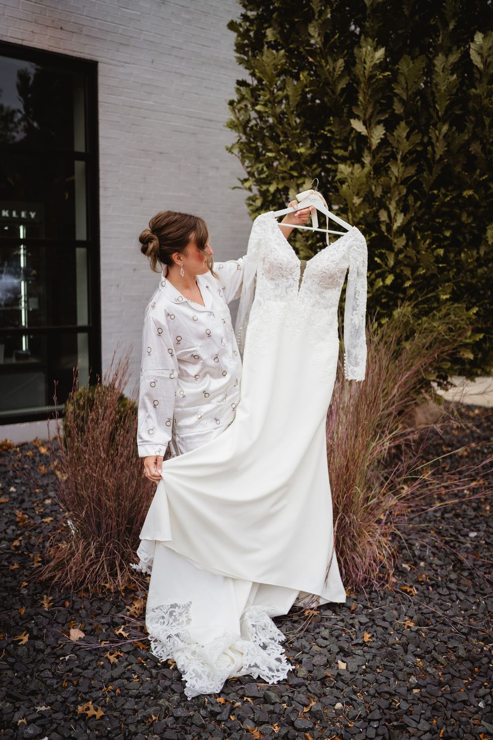A wedding dress with lace details and long sleeves is displayed against a white brick wall and green foliage.