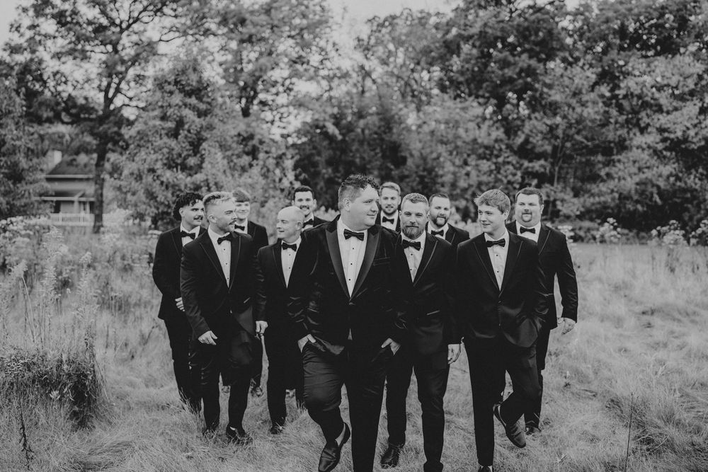 Groomsmen in black suits walk together outdoors through a garden setting.