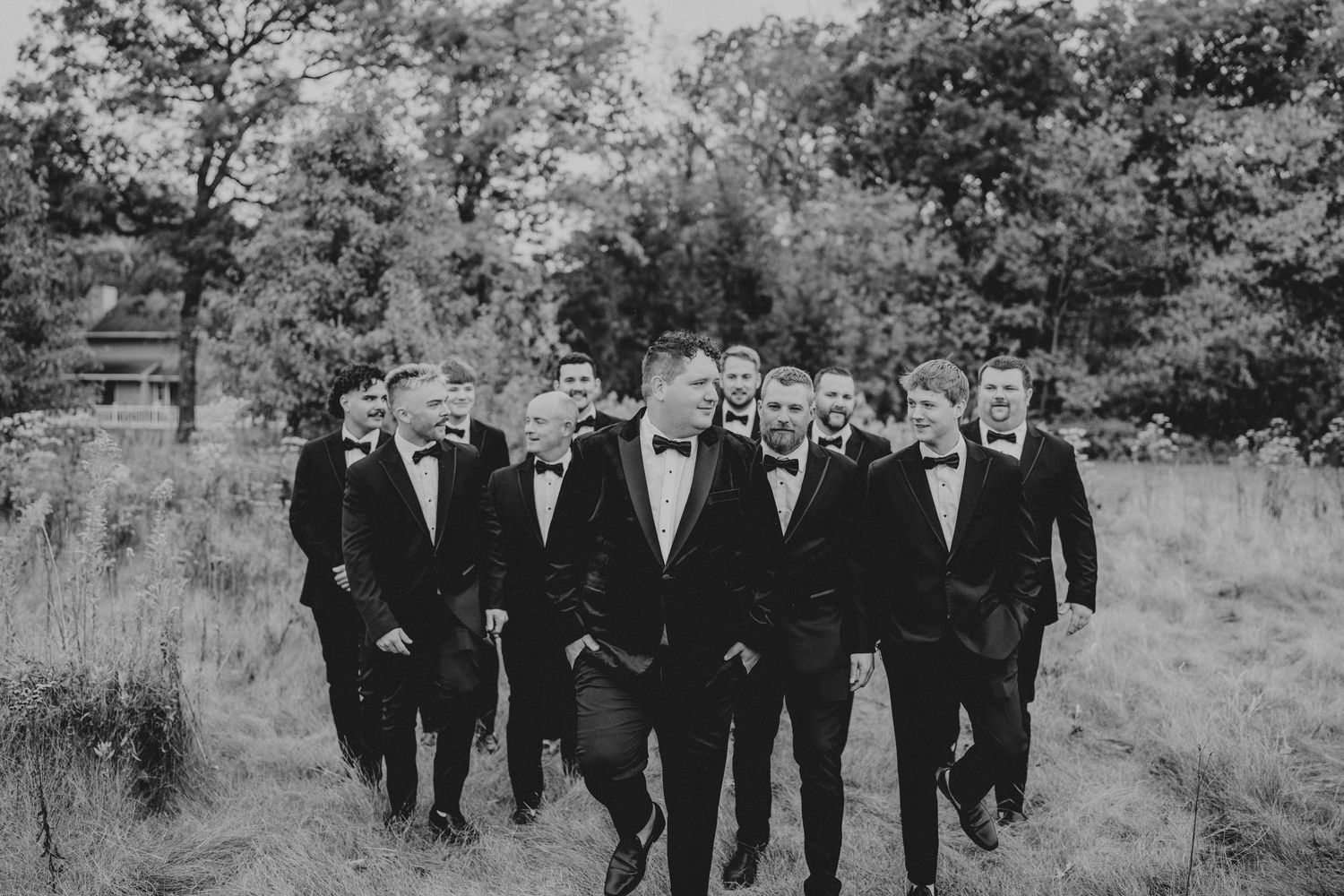 Groomsmen in black suits walk together outdoors through a garden setting.