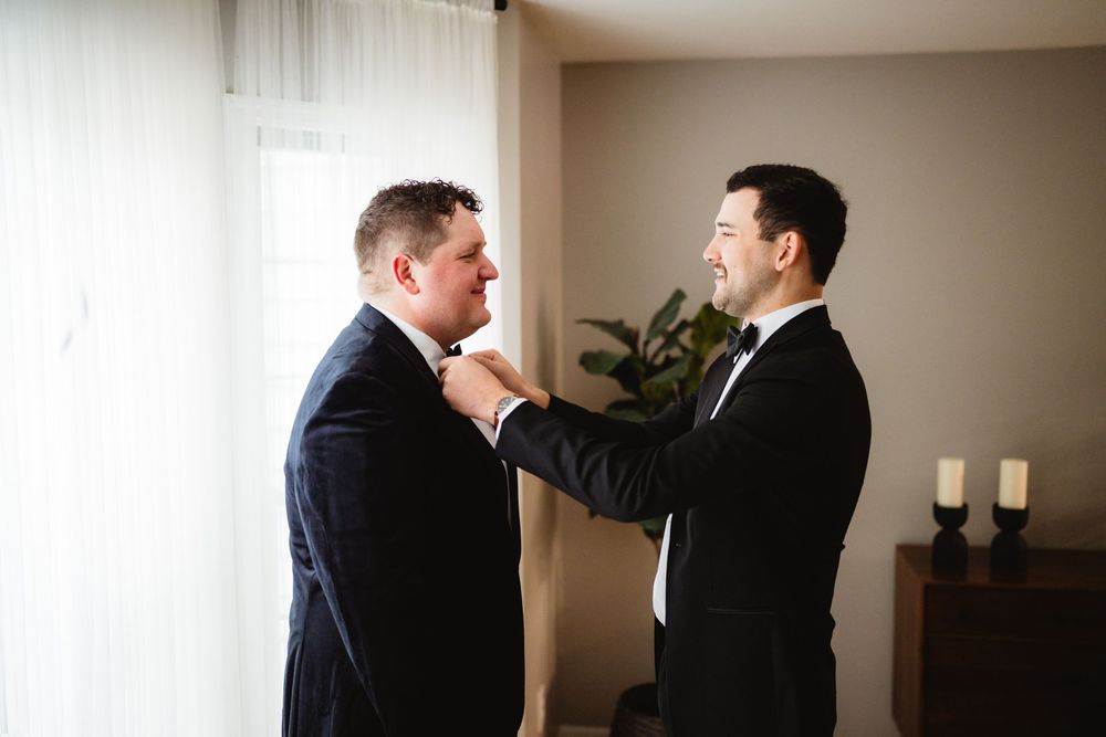 Two people in dark suits help each other adjust their ties while getting ready in a room with natural lighting.