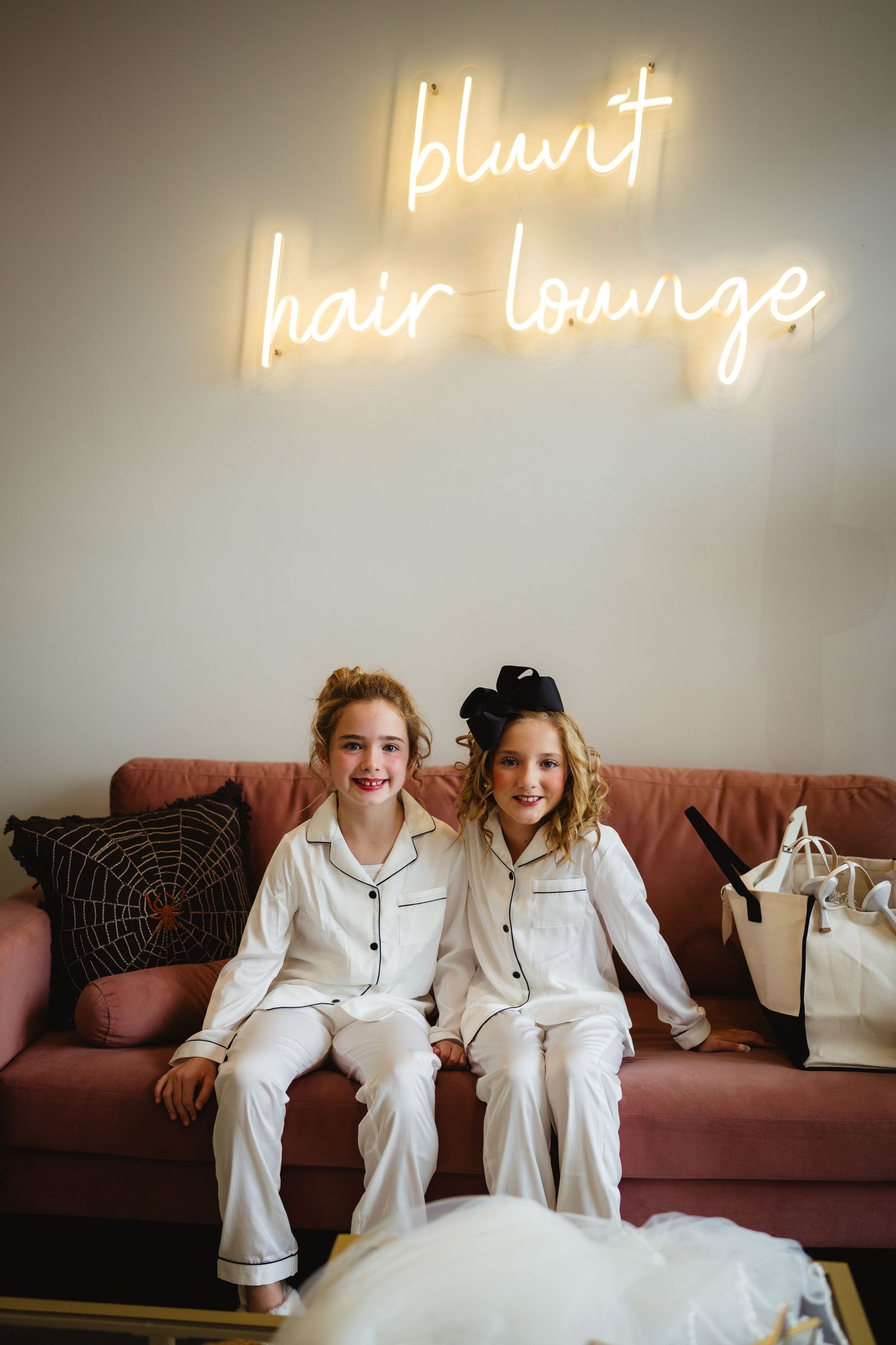 Two children in matching white pajamas sit on a pink couch beneath illuminated 'blunt hair lounge' neon signage.