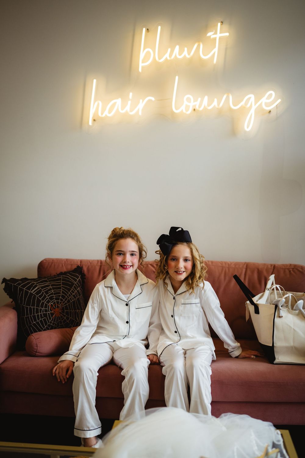 Two children in matching white pajamas sit on a pink couch beneath illuminated 'blunt hair lounge' neon signage.