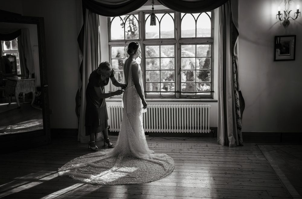 A black and white photo of a wedding dress fitting in an elegant room with gothic windows and hardwood floors.