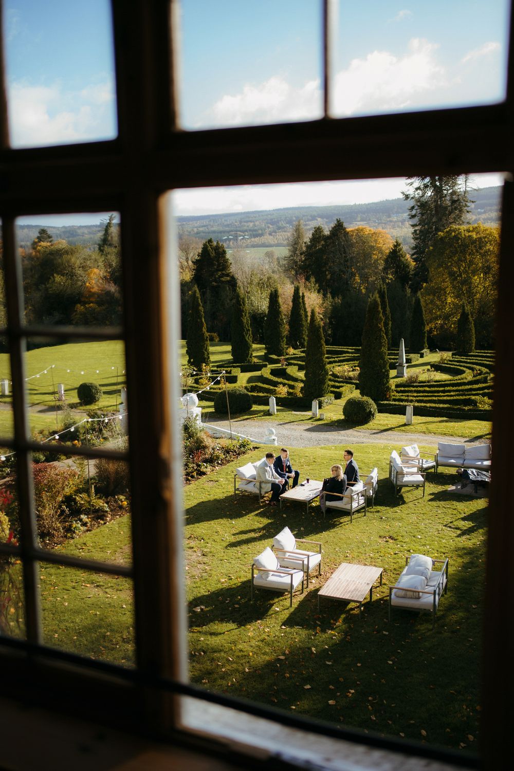 View through a window of outdoor lounge chairs arranged on a manicured garden lawn with formal hedges and trees.