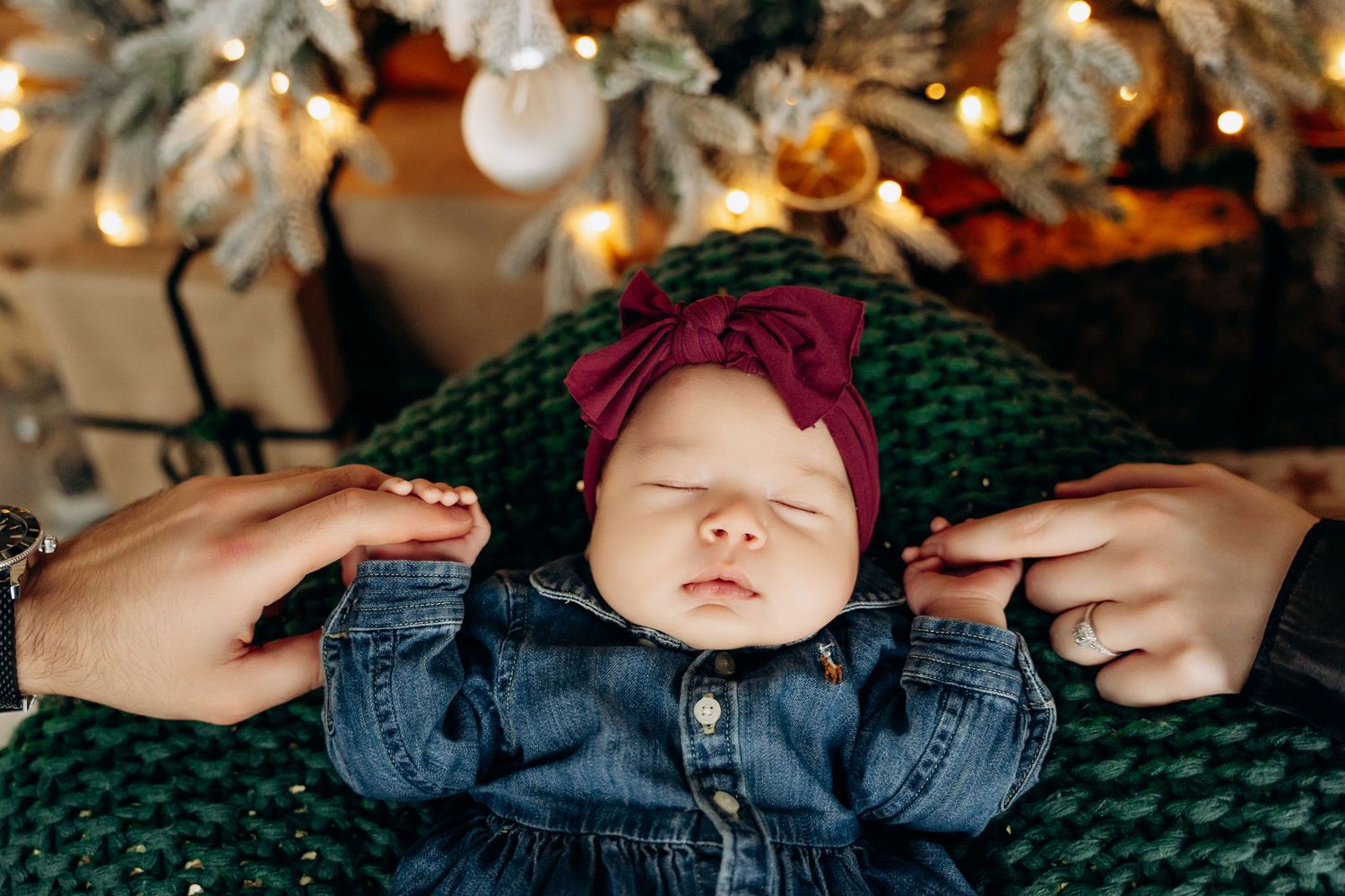 A sleeping baby wearing denim and a burgundy headband rests on a green blanket beneath Christmas decorations.