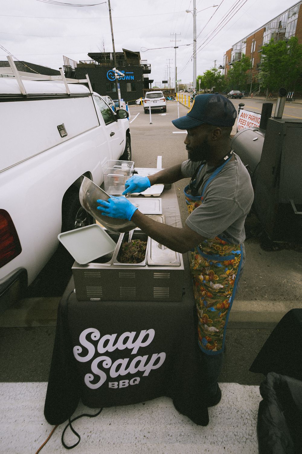 Man in blue gloves preparing food at Saap Saap BBQ outdoor street food stand next to a white van.
