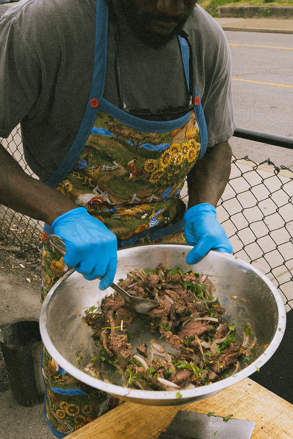 Man in floral apron and blue gloves preparing seasoned crab in a large metal bowl outdoors.