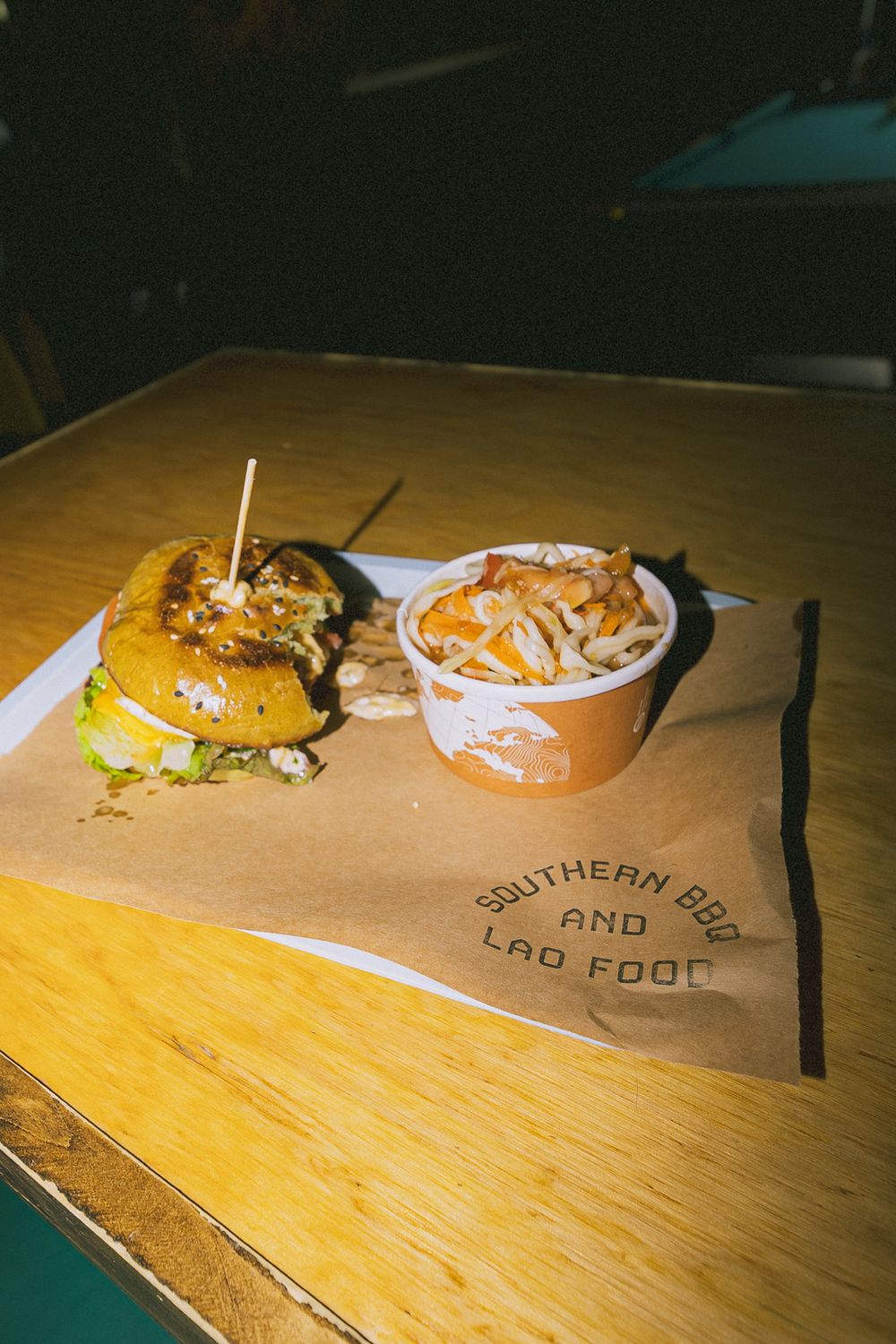 A burger and coleslaw side on Southern BBQ and Lao Food branded paper at a wooden table.