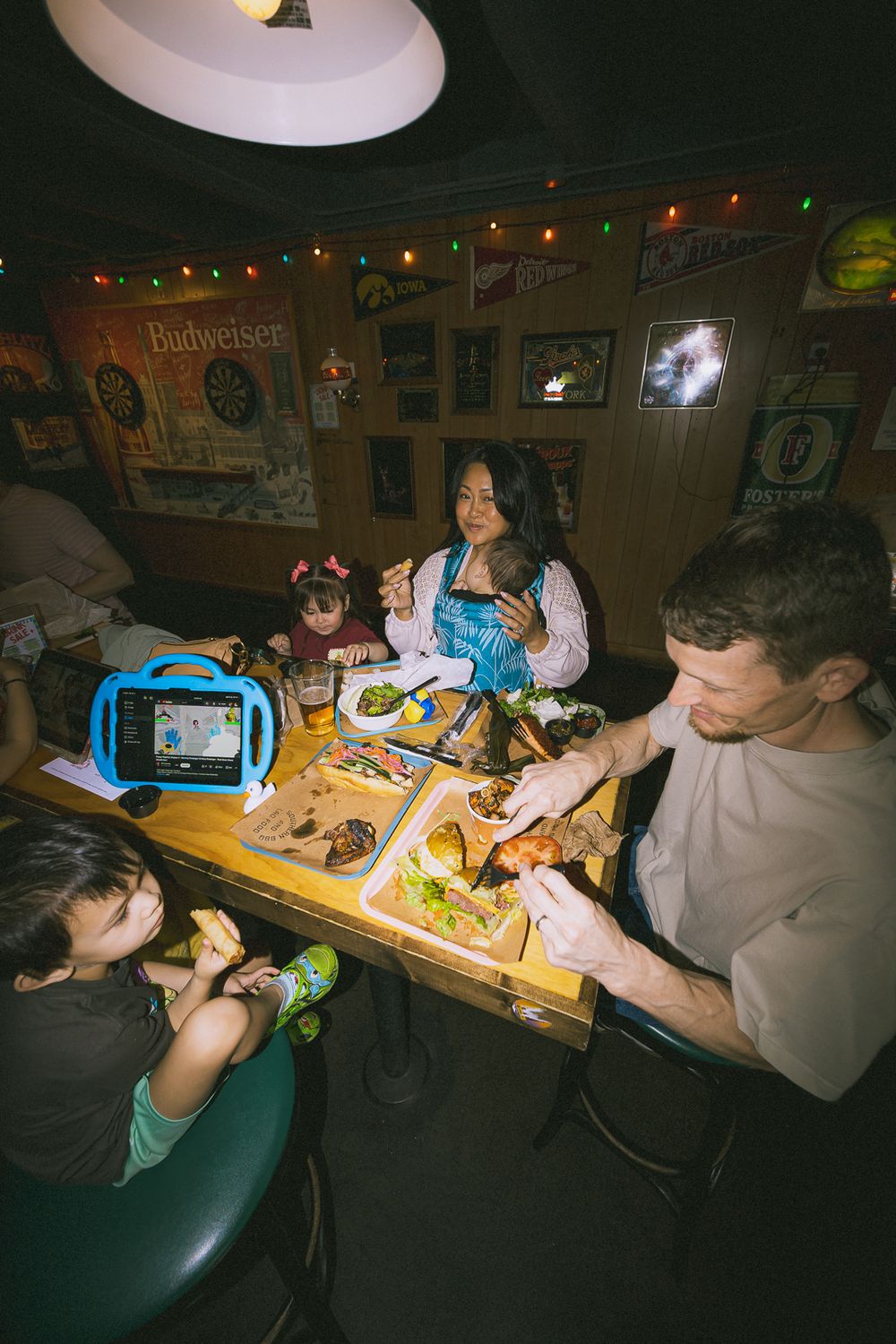 Family dining at a casual bar restaurant with kids, food trays, and a tablet on the table under warm string lights.