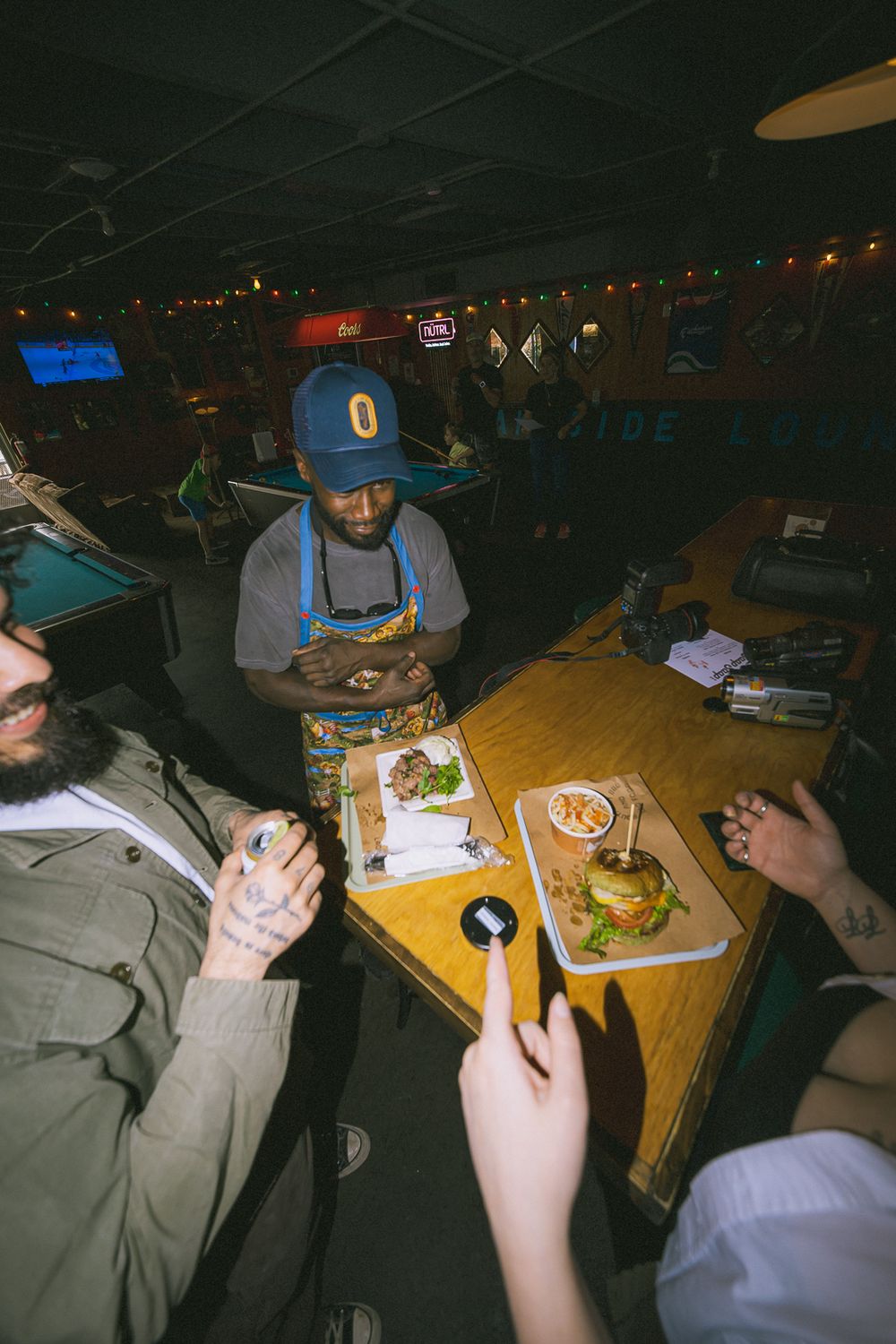 People enjoying burgers at a casual bar restaurant, with a server in a blue cap delivering food trays to the table.