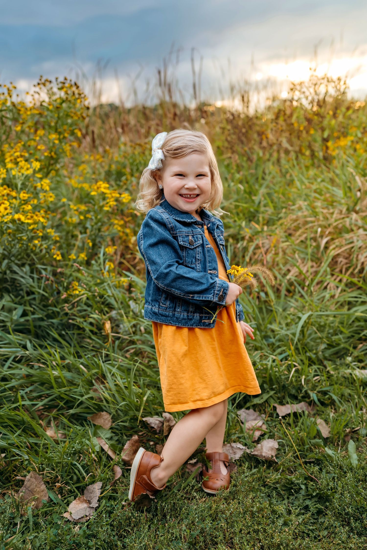 Child wearing denim jacket and orange dress plays joyfully in field of wildflowers.