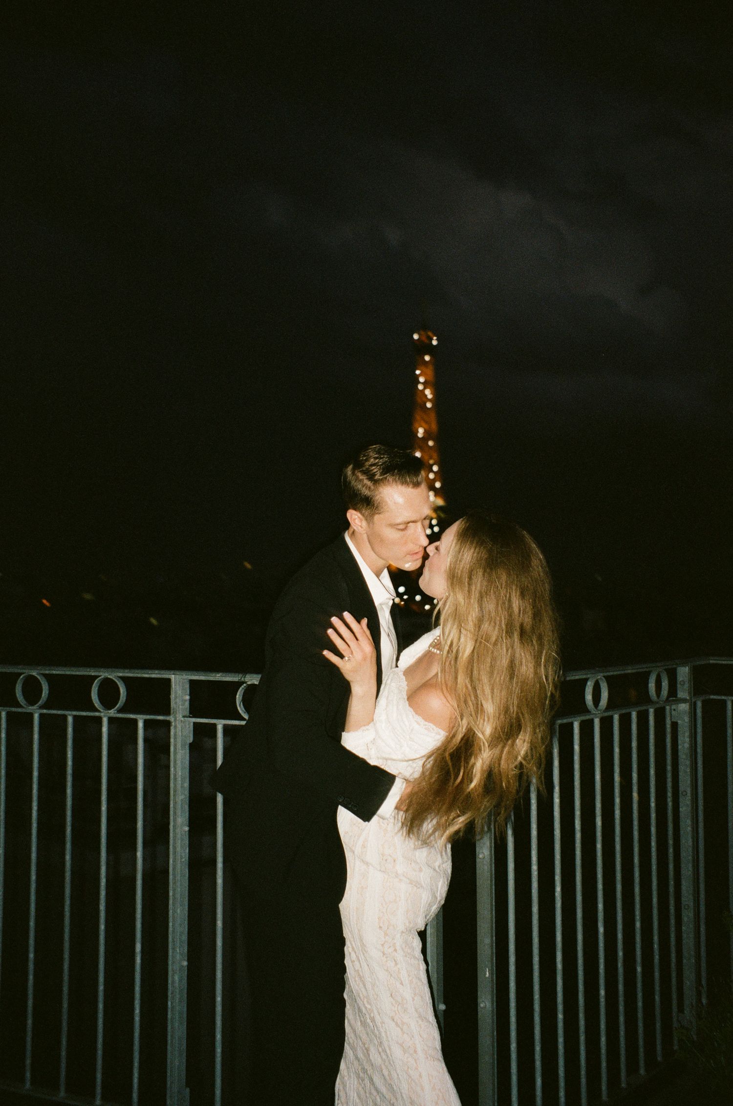 A couple in wedding attire share romantic kisses on a balcony at night with twinkling lights in the background.