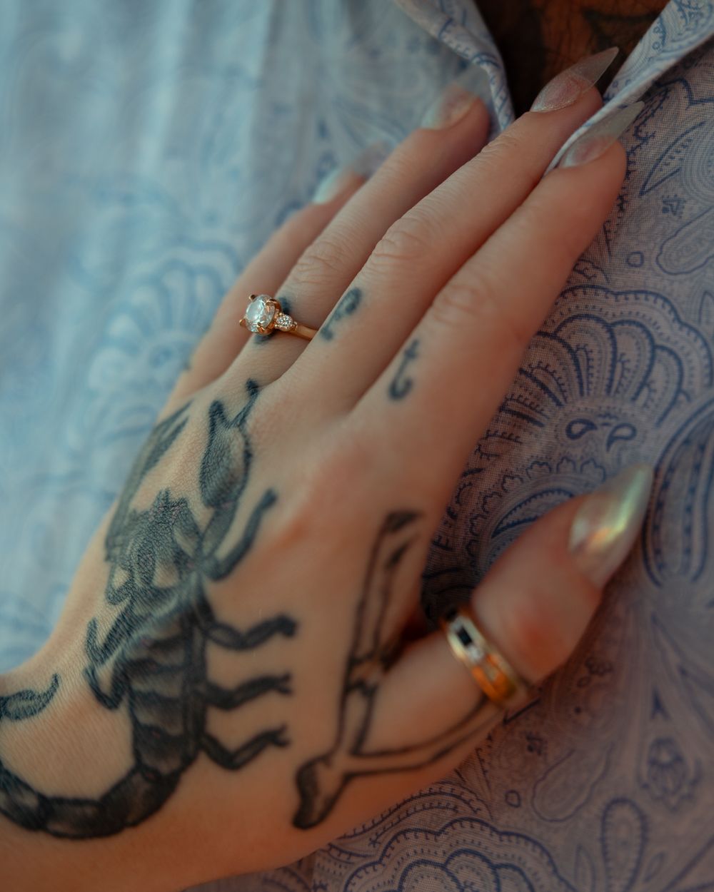 Hand with a detailed scorpion tattoo wearing two rings, including an engagement ring with a diamond solitaire.