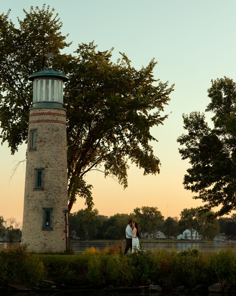 Stone lighthouse tower stands against a sunset sky with trees silhouetted in the background at dusk.