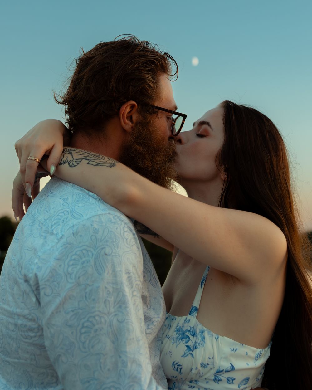 Romantic couple sharing an intimate moment at sunset with the moon visible in a turquoise sky.