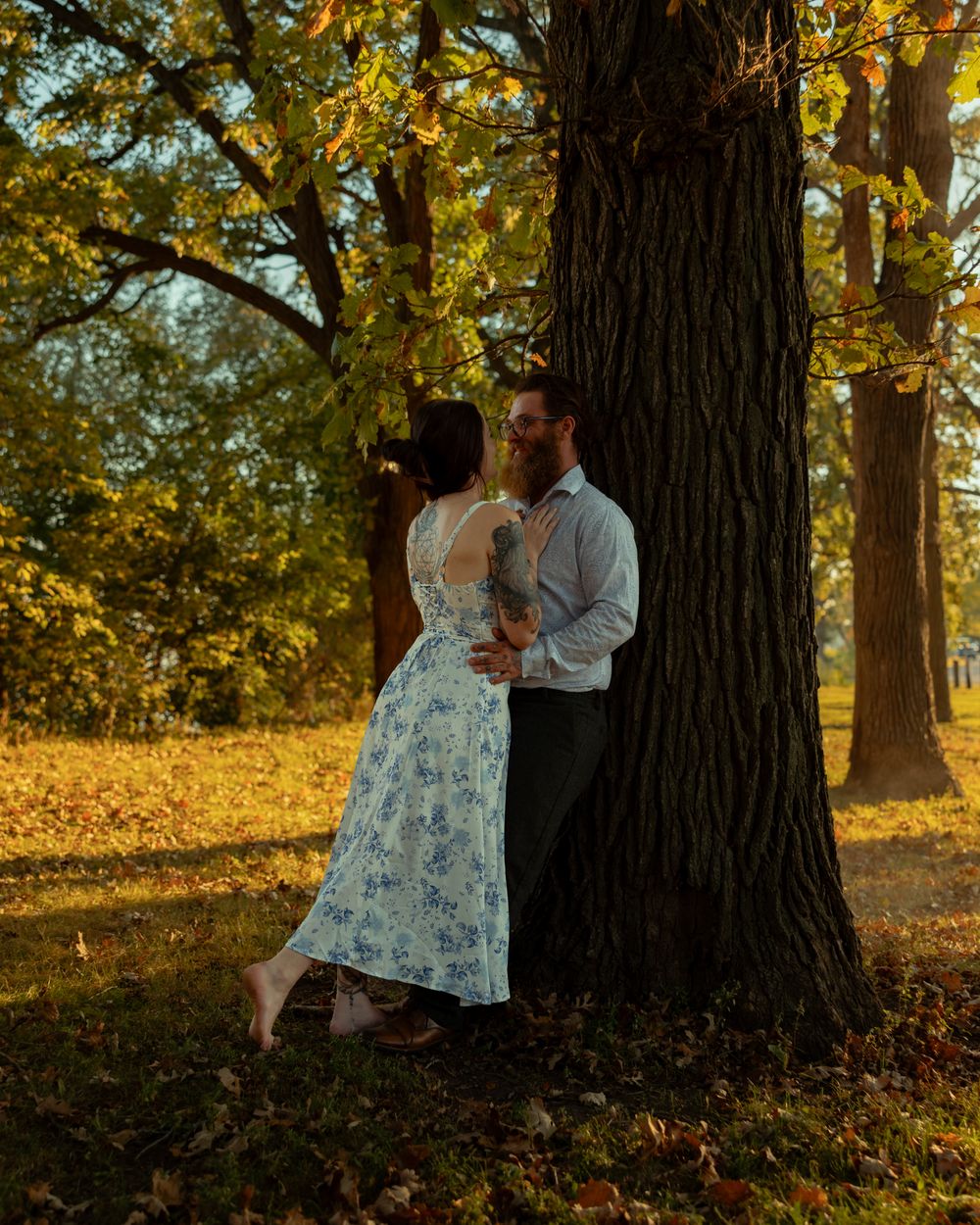 A couple shares an intimate moment against a large tree trunk in a sunlit autumn park setting.