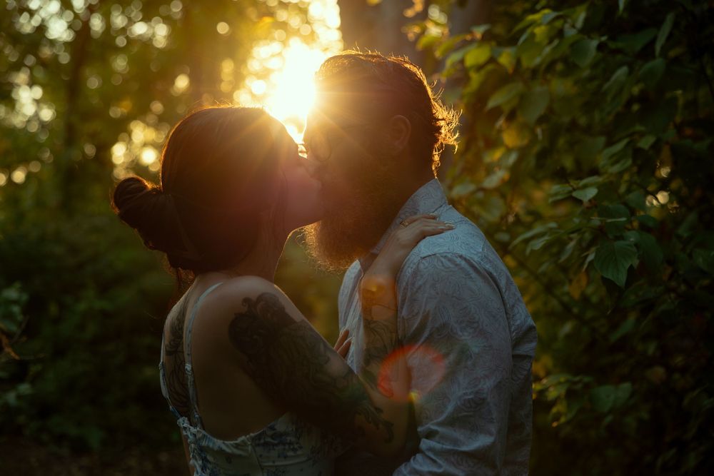 A silhouetted couple shares a romantic kiss at sunset with golden sunlight streaming through trees in the background.