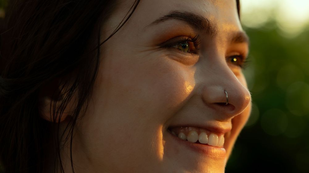 Side profile portrait of a smiling person with a nose piercing in warm golden sunlight.