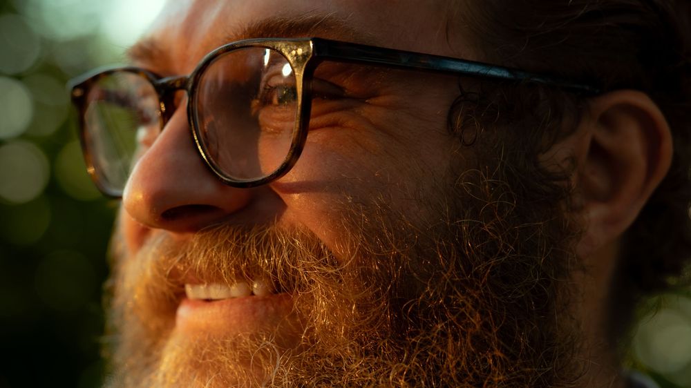 Close-up view of a red beard and glasses against a blurred natural background.