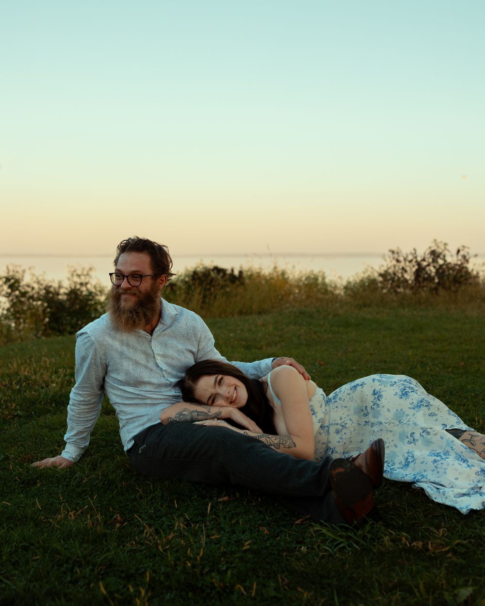 A couple relaxes on a grassy hillside at sunset, with one person resting their head on the other's lap.