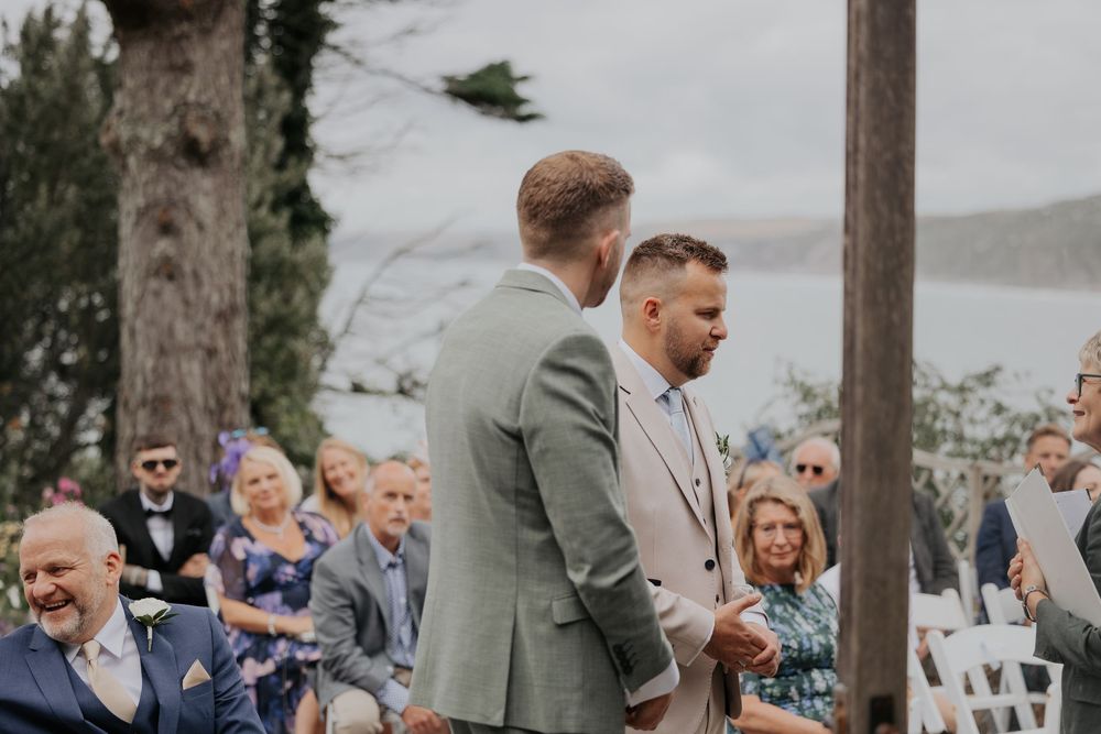 Groom in beige suit standing at outdoor wedding altar with best man in sage green suit.