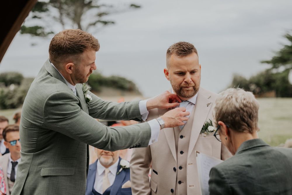 Best man in sage green suit adjusting groom's boutonniere at an outdoor wedding ceremony.