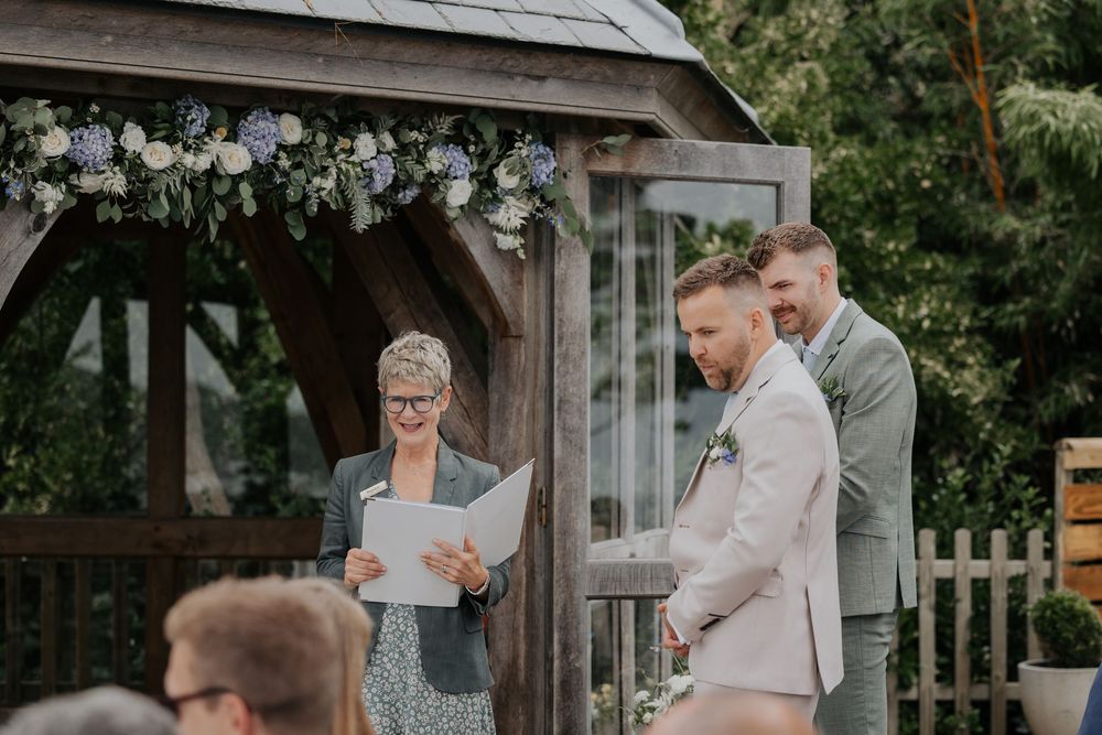 Groom reads vows at outdoor wedding ceremony beneath floral-adorned wooden gazebo with officiant.