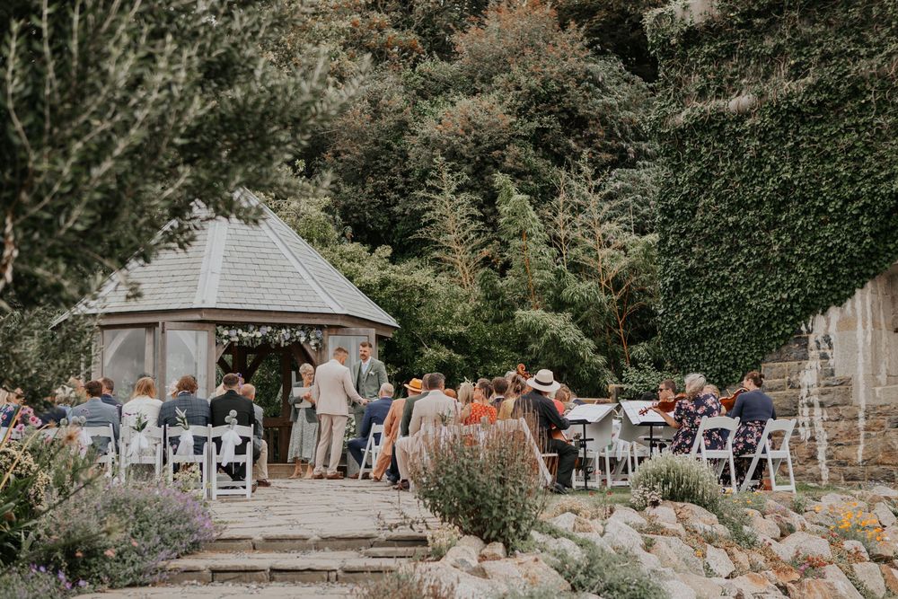 Guests seated outdoors at elegant garden wedding reception near rustic wooden gazebo.