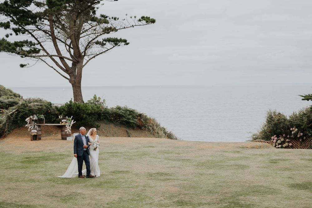 Bride and groom stand together on coastal clifftop lawn with ocean backdrop.