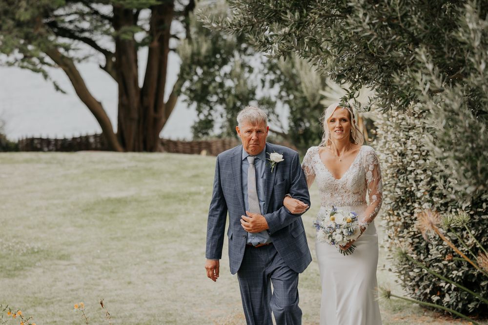 Father walks bride down the aisle through lush garden setting on wedding day.