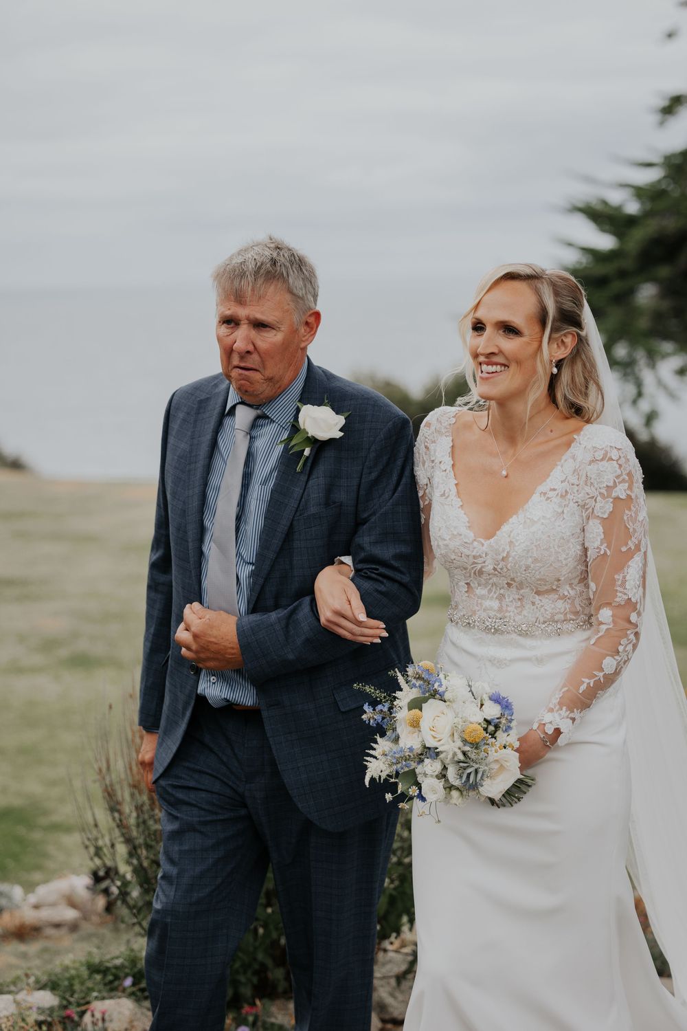 Smiling bride in lace gown walks with father in blue suit holding bouquet.