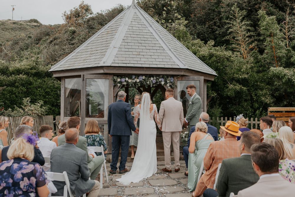 Bride and groom exchange vows at wooden gazebo altar before seated wedding guests.