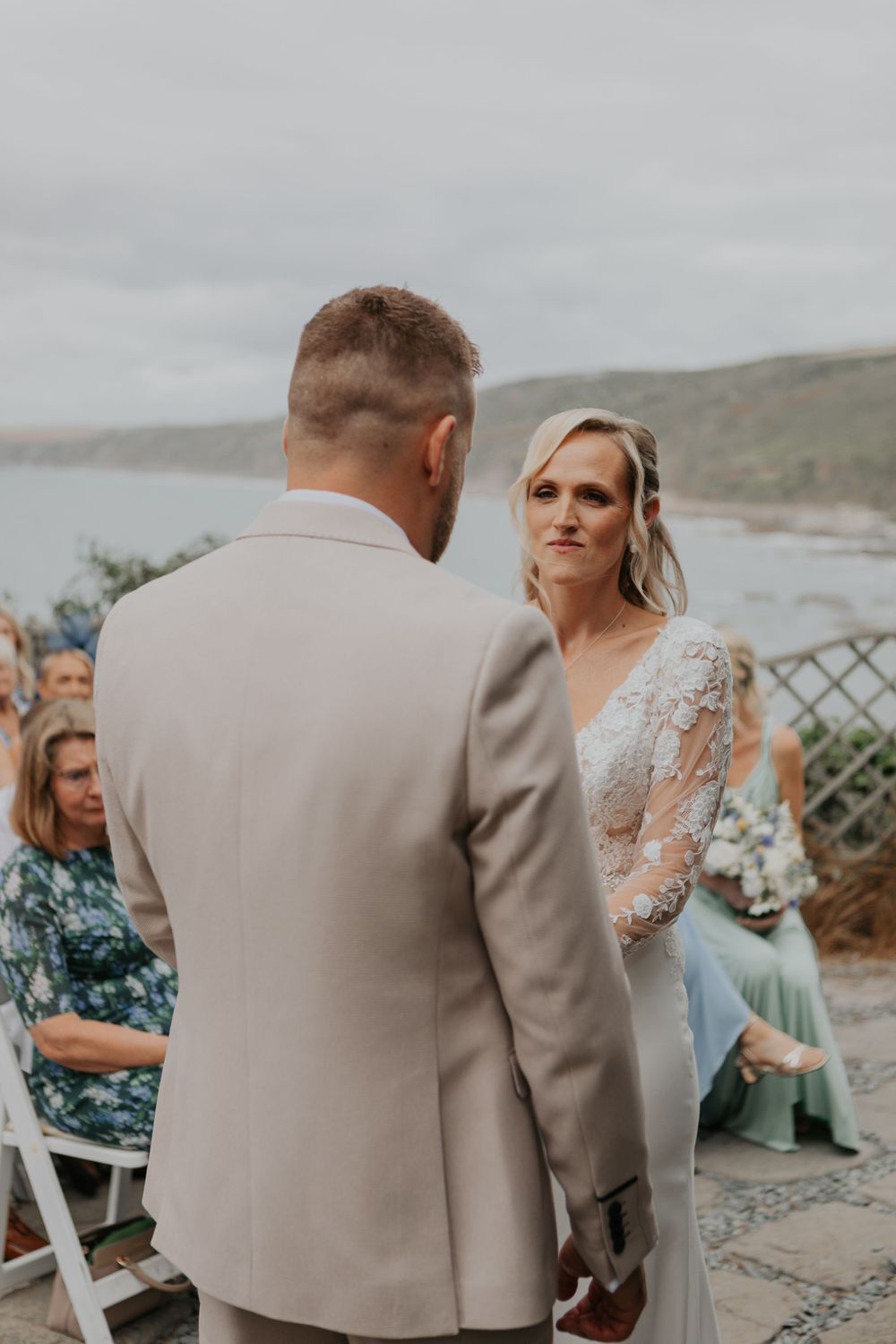 Emotional bride gazes at groom during outdoor coastal wedding ceremony with guests.