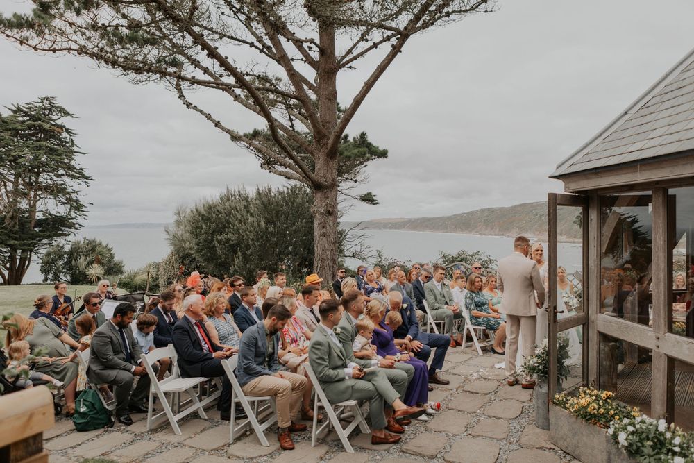 Wedding guests seated outdoors overlooking coastal scenery during ceremony by wooden gazebo.