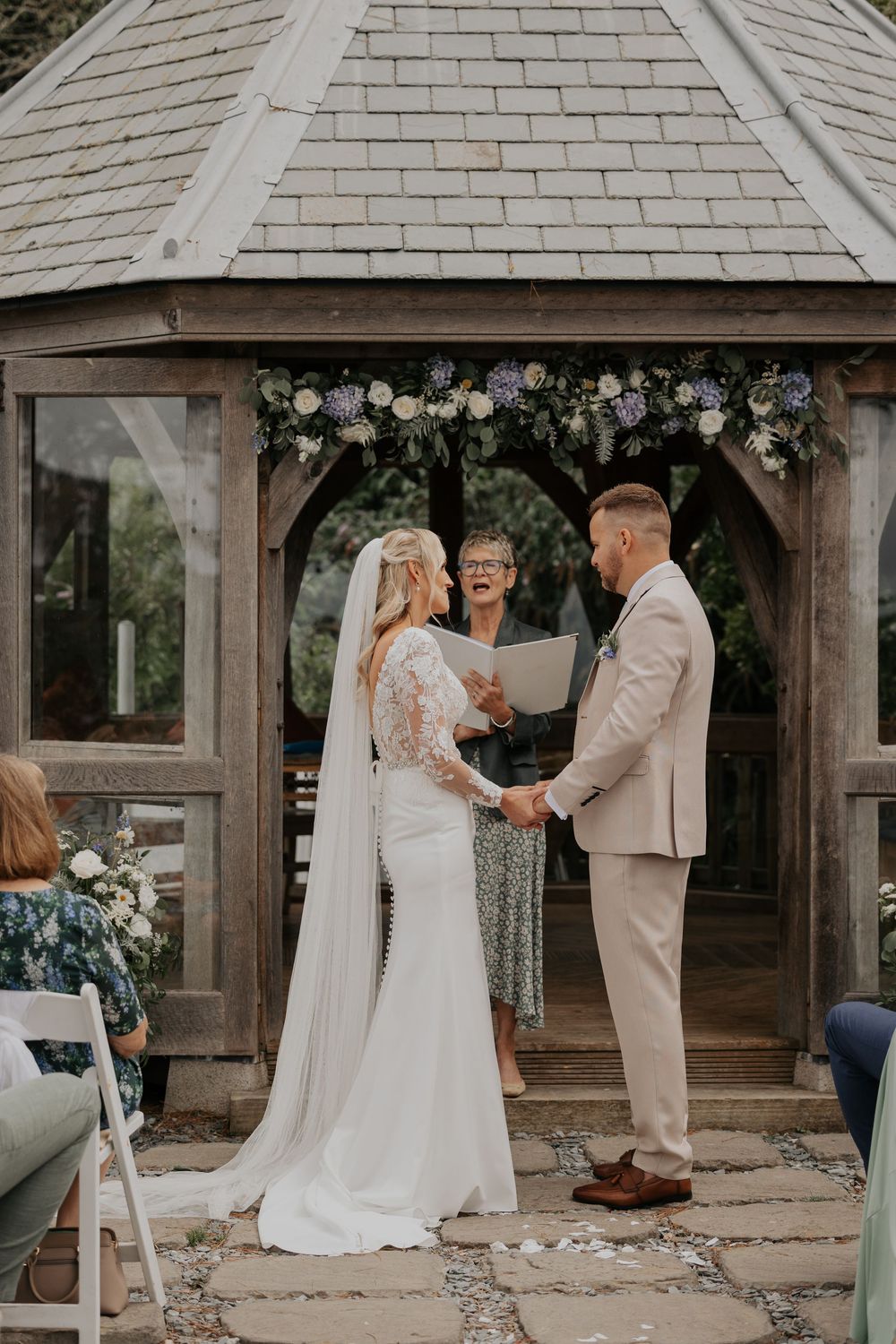 Bride in lace gown and groom hold hands at decorated gazebo wedding ceremony.