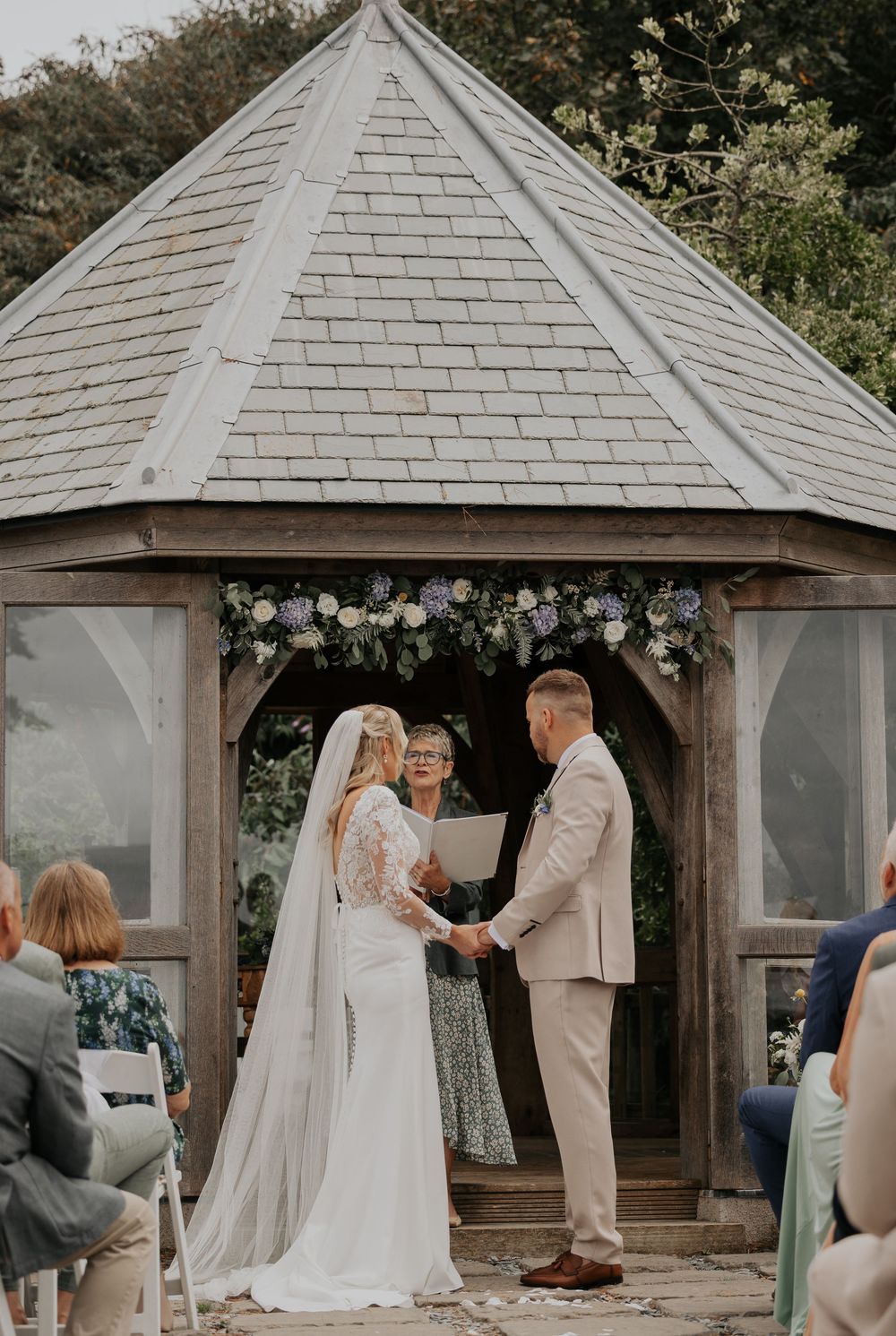 Bride and groom hold hands at floral gazebo altar with officiant during ceremony.