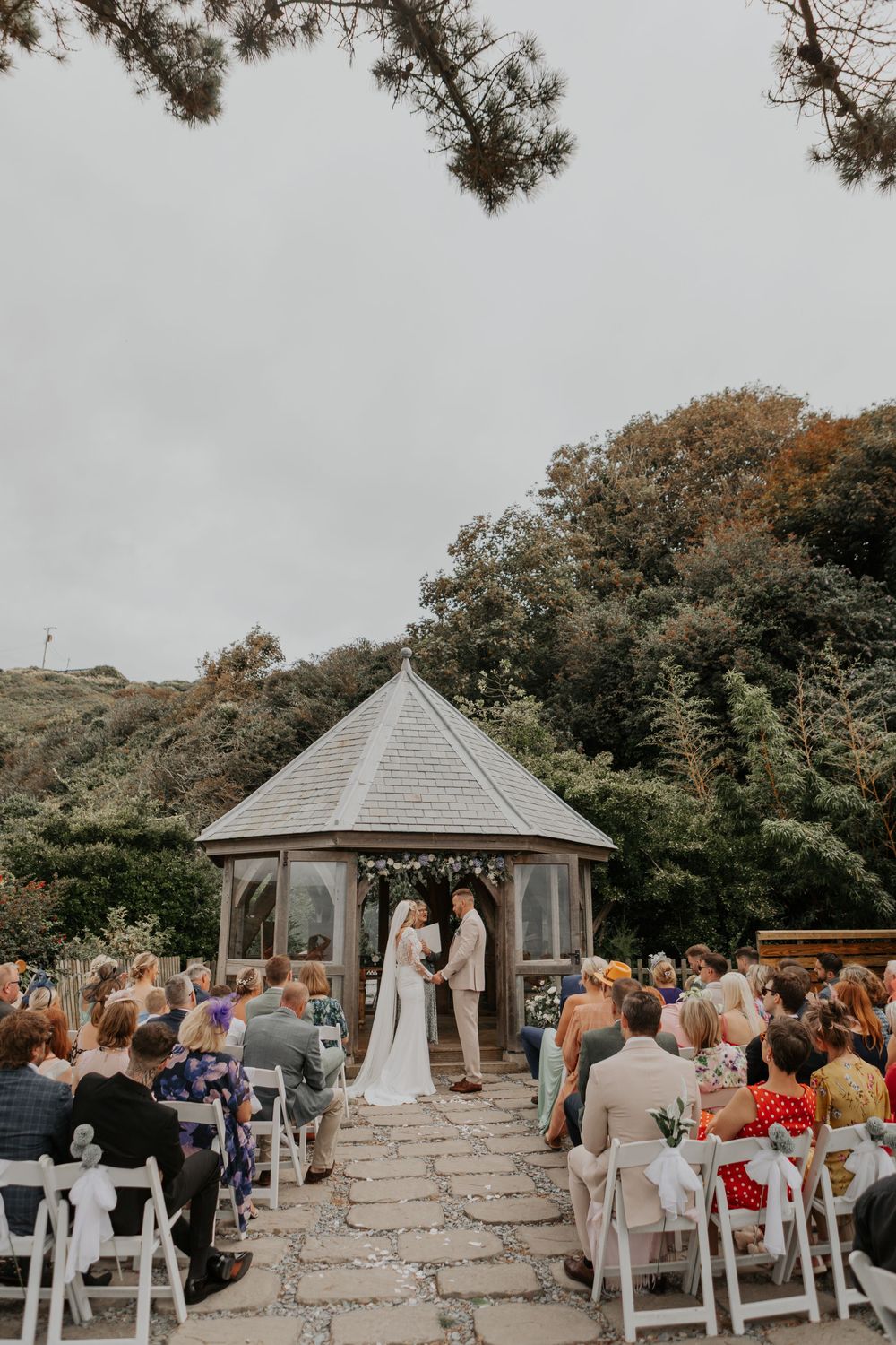Outdoor wedding ceremony at a gazebo with guests seated on white chairs along a stone path.