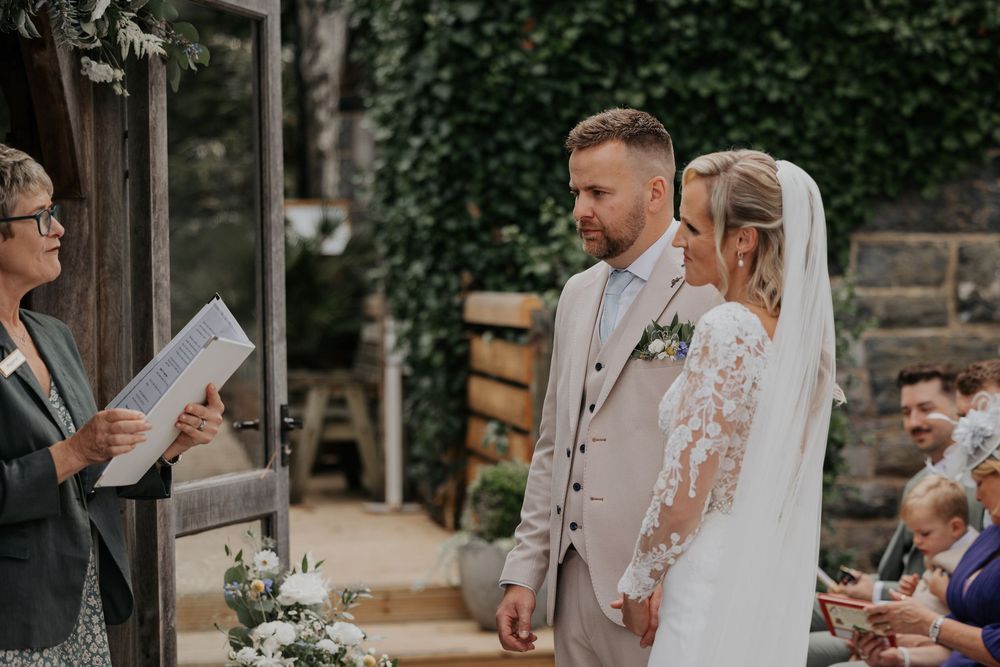 Officiant reads vows as groom in beige suit and bride in lace dress stand at the altar.