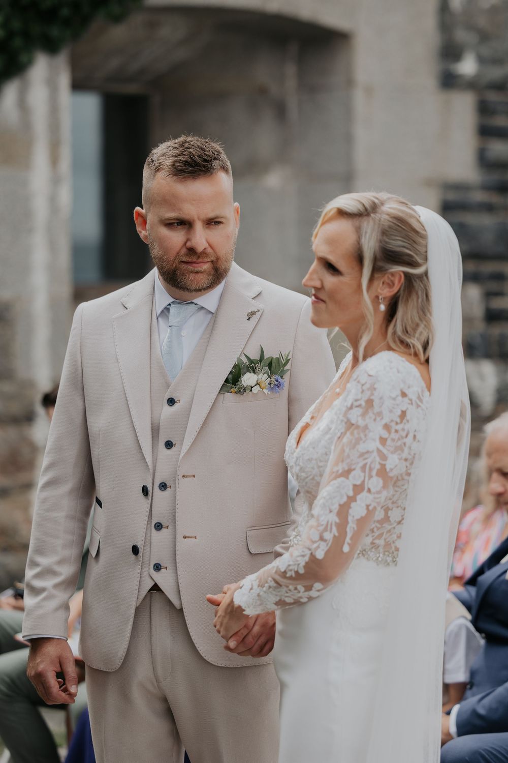 Groom in tan suit holds bride's hands during outdoor wedding ceremony at stone venue.